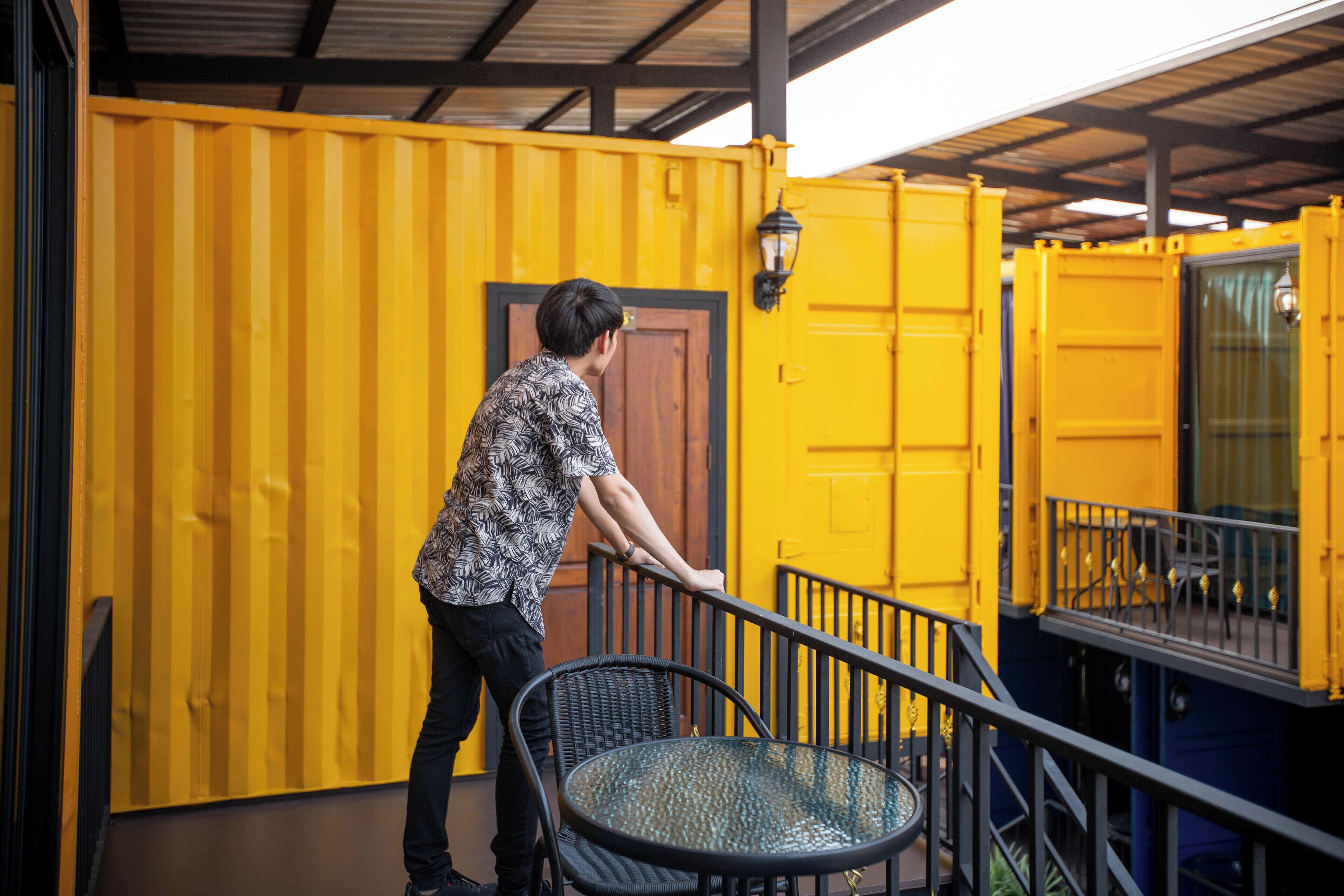 A person leaning on a railing next to a yellow shipping container building with a door and a window.