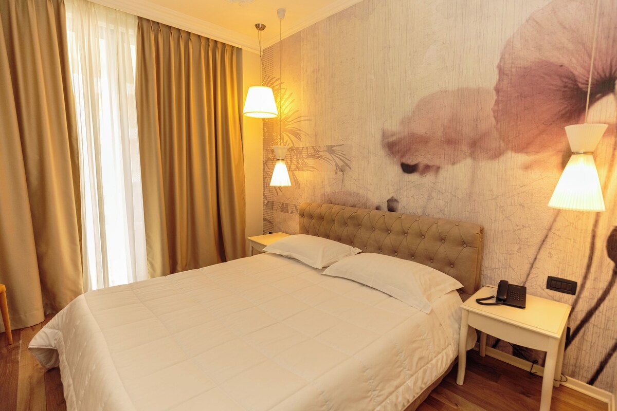 A hotel room with a large bed, two bedside lamps, a nightstand with a phone, and a wall with a floral mural.