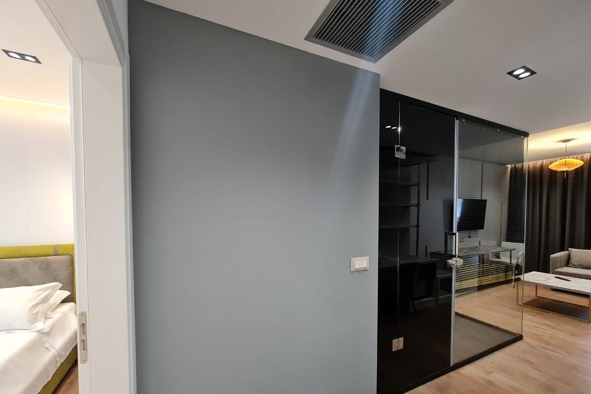 A modern hotel room with a bed, a glass partition, a TV, and a dining area.