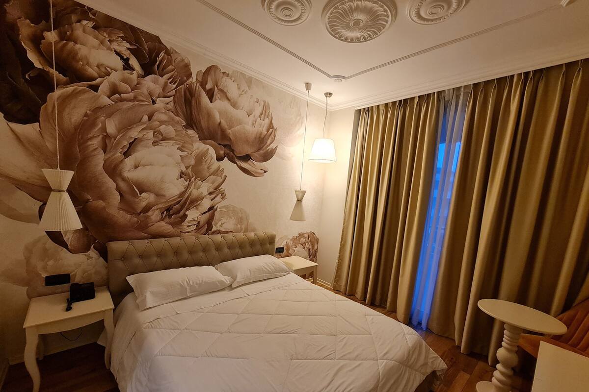 Une chambre avec un grand lit, une table de chevet, une lampe et un mur orné d’une fresque florale.