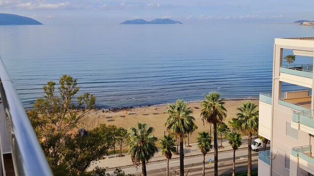 A coastal view with a beach, palm trees, and a road leading to a building with balconies overlooking the sea. - Regina City Hotel (Vlorë)