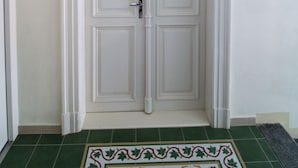 Une porte double blanche, ornée d’une poignée de porte dorée et encadrée de moulures décoratives, se trouve dans un couloir dont le sol est recouvert de carreaux verts et où repose un tapis à motifs.