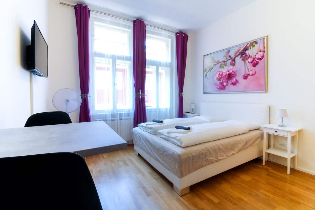 Habitación de hotel con cama, escritorio, silla, mesita de noche con lámpara y una ventana grande con cortinas.