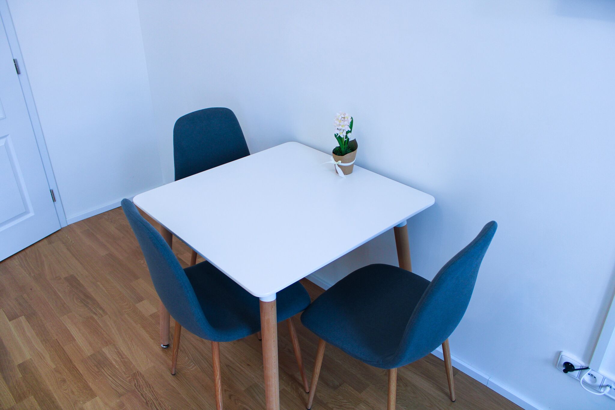 Un pequeño comedor con una mesa blanca y dos sillas azules, una planta en maceta sobre la mesa, y un piso de madera.