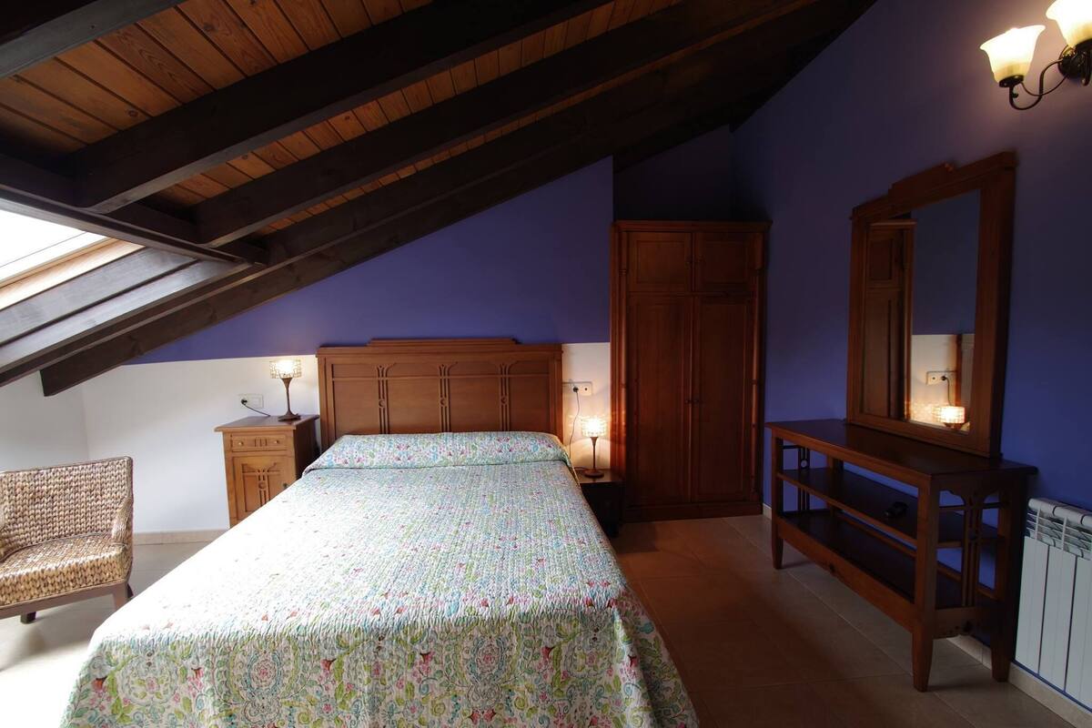 A bedroom with a sloped ceiling, a bed with a patterned bedspread, a wooden dresser, a nightstand with a lamp, a chair, and a large mirror.