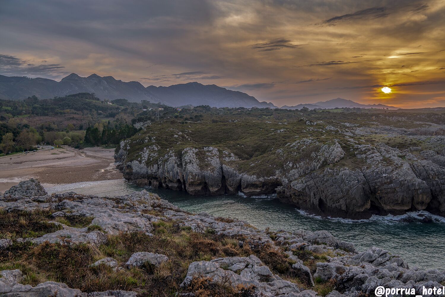 Eine Küstenlandschaft mit felsigem Gelände, einem Fluss und entfernten Bergen unter einem Sonnenuntergangshimmel.