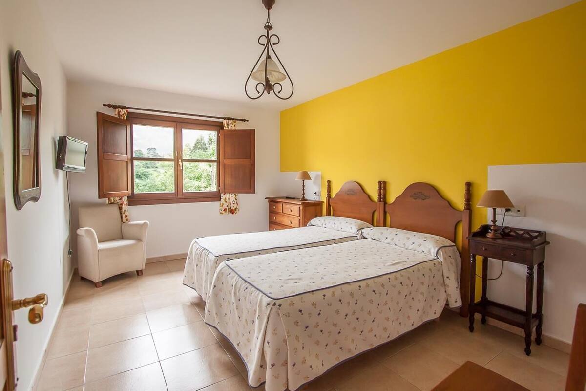 A bedroom with two beds, a chair, a bedside table, a lamp, and a window with curtains.