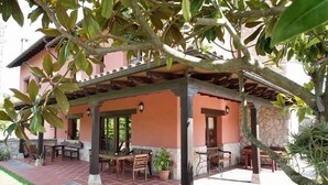 A covered outdoor seating area with tables and chairs, a stone wall, and a building with a tiled roof in the background. - Hotel Rural Porrua (Llanes)