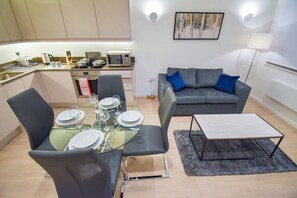 A modern living room with a grey sofa, a glass dining table set for four, and a kitchen in the background.