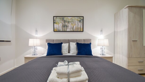 A neatly made bed with a grey quilt, two blue pillows, and a stack of white towels tied with a ribbon. - Silbury MK City Apartment With Parking (Milton Keynes)