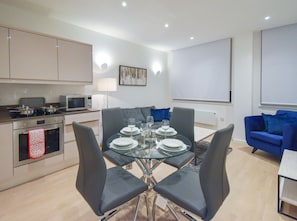 A modern kitchen with a dining area, including a glass table set for six, grey upholstered chairs, a blue sofa, and a flat-screen TV mounted on the wall.