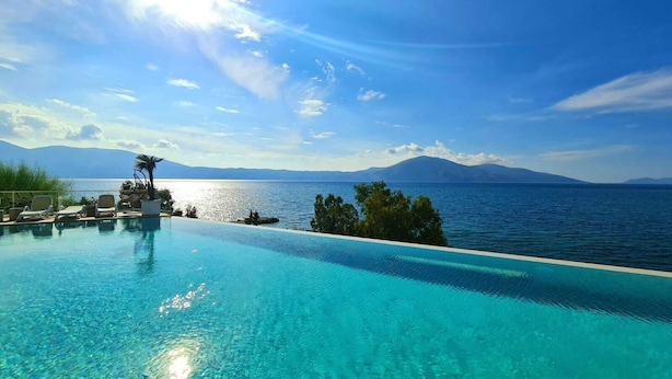 Ein Pool im Überlaufstil mit klarem, blauem Wasser, Liegestühlen und Blick auf das Meer und die Berge.