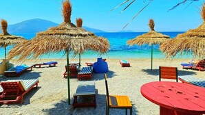 Ein Strand mit reetgedeckten Sonnenschirmen, Liegestühlen und einem Tisch.