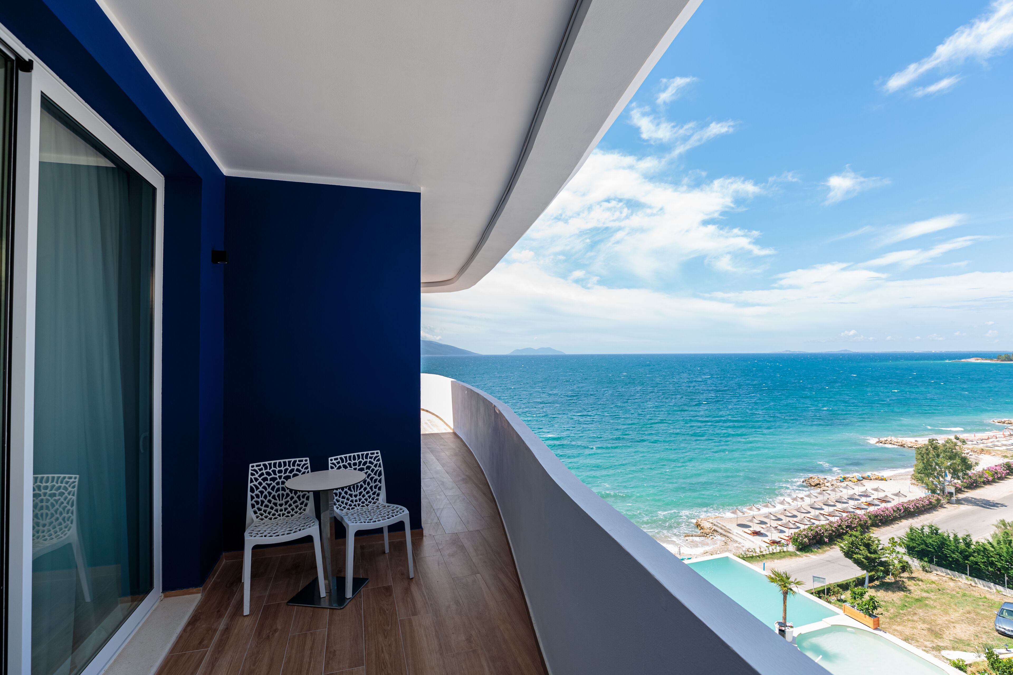 A balcony with a view of the sea, a pool, and beach umbrellas.