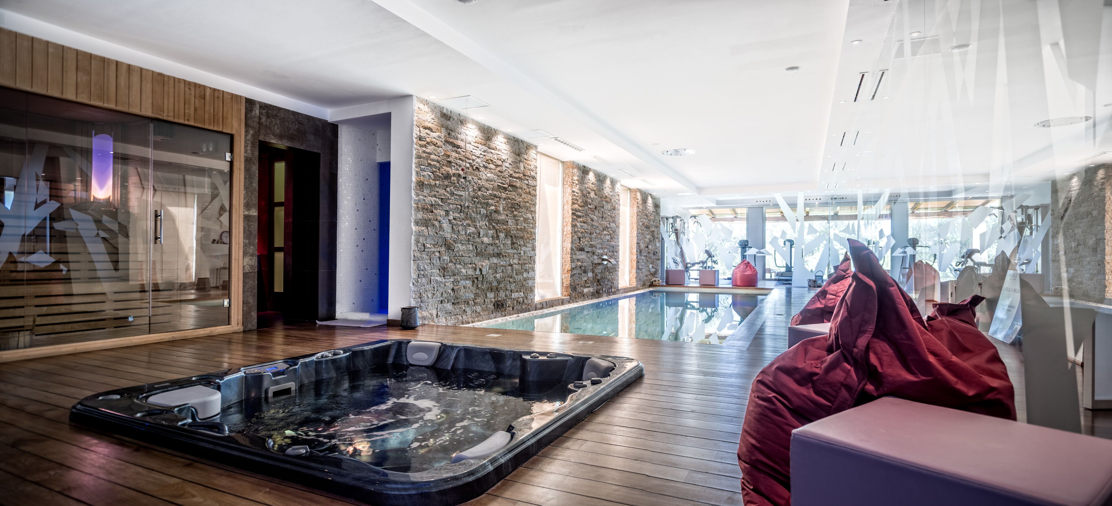 A modern spa area with a hot tub, wooden flooring, stone walls, and a glass-enclosed pool.
