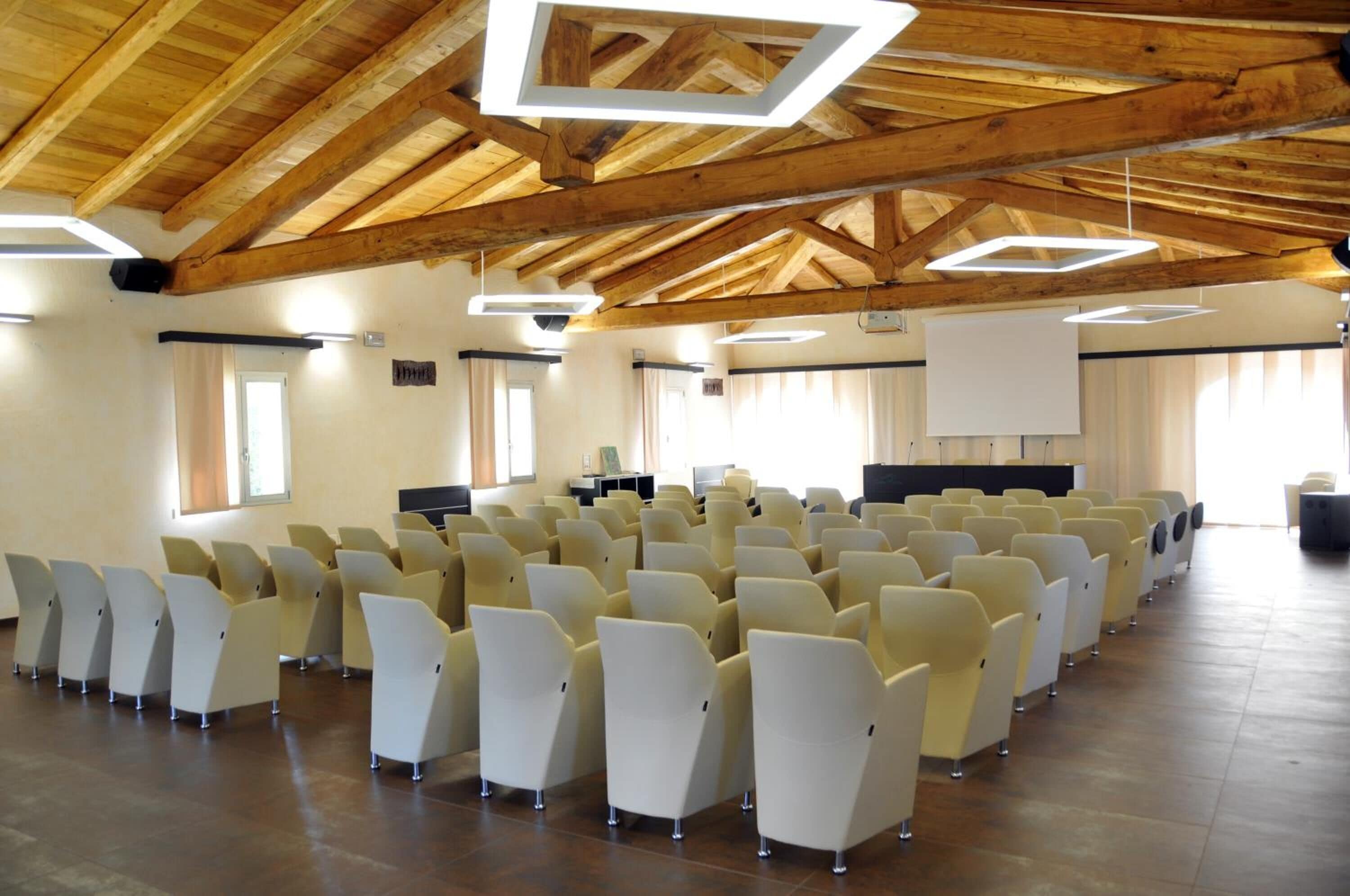 Un salón de conferencias con filas de sillas, un escenario con una pantalla y vigas de madera en el techo.