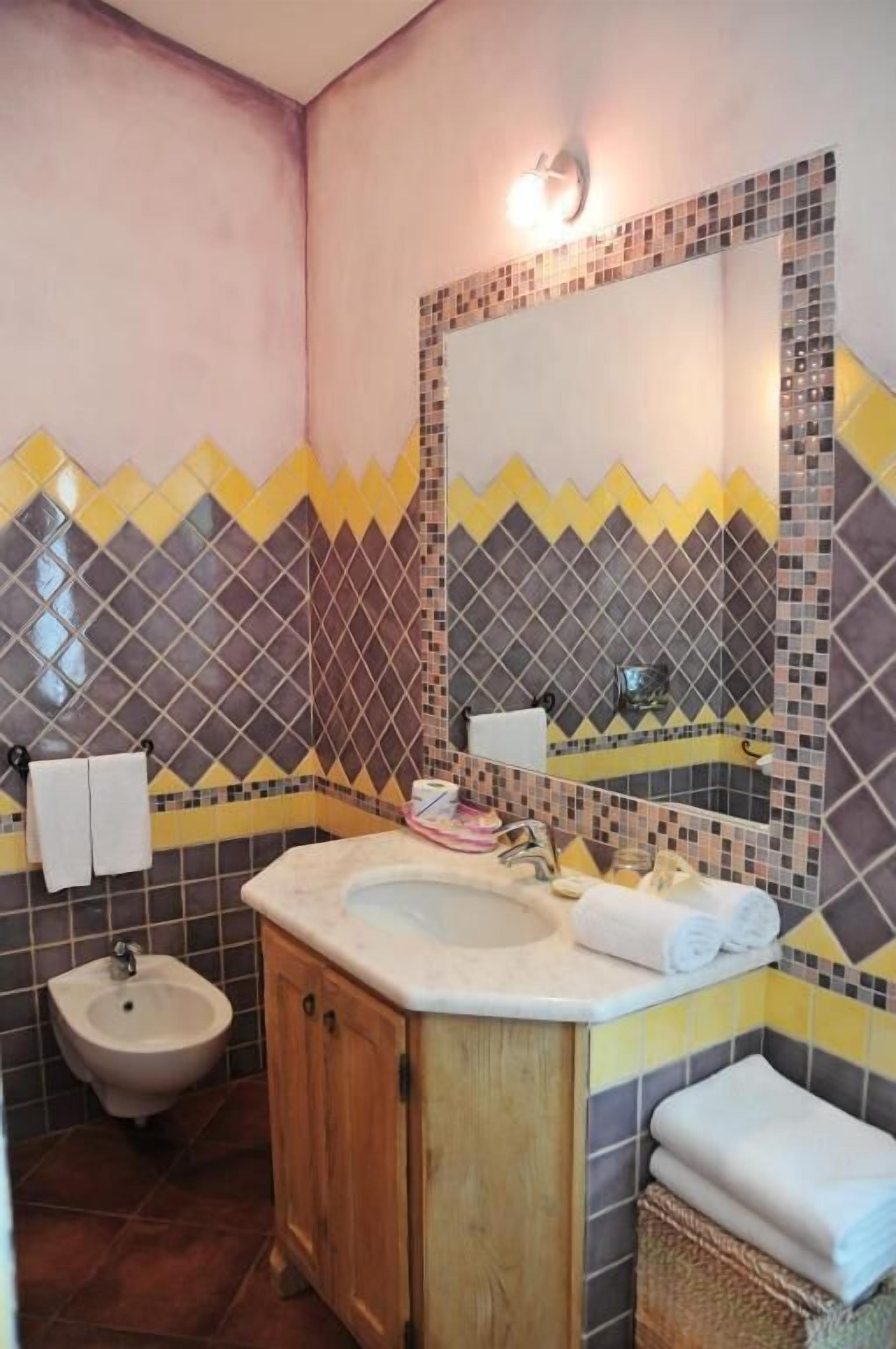 Een badkamer met een wastafel, spiegel en bidet. De wandtegels hebben een zigzagpatroon in geel en grijs.