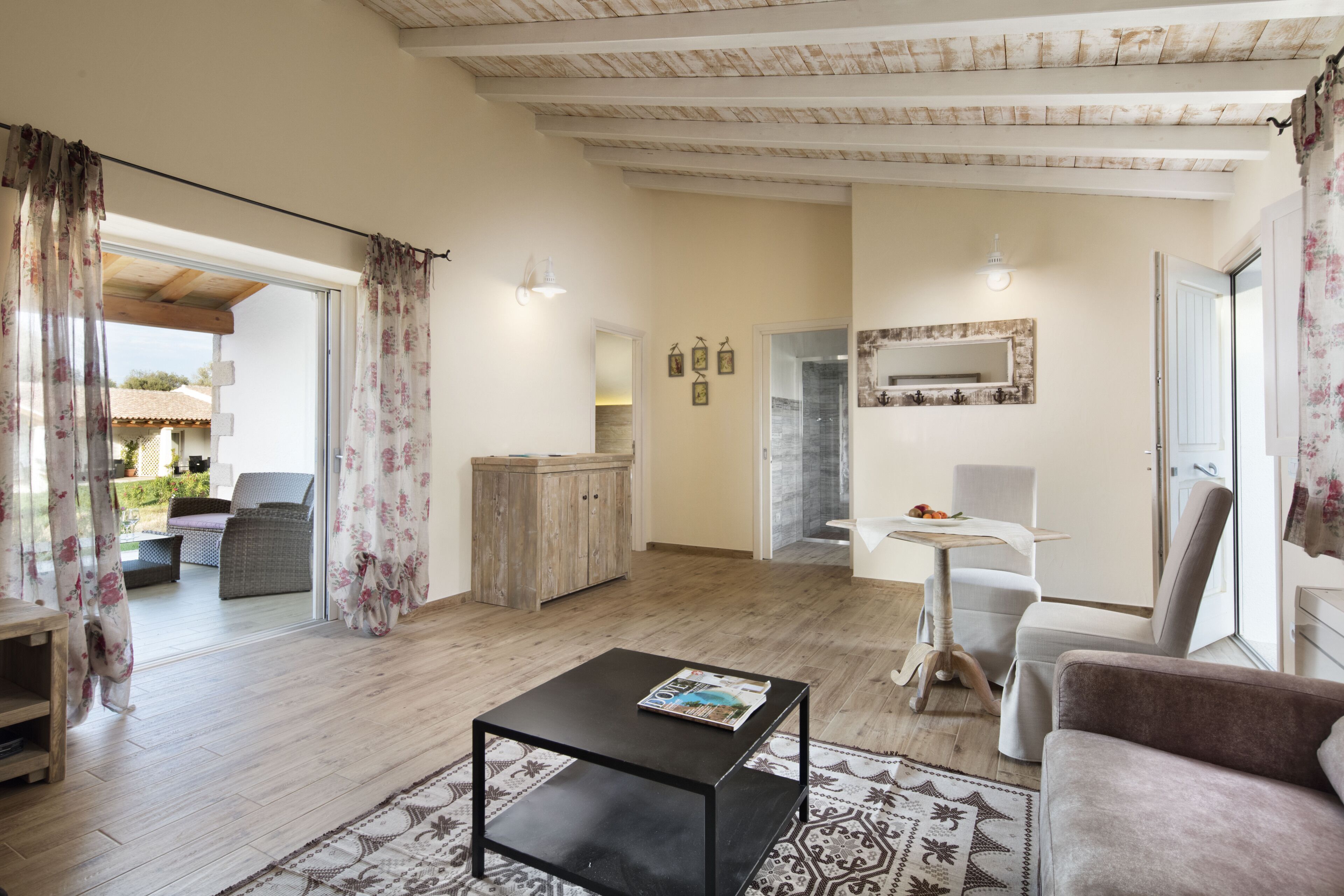 A spacious living area with a wooden floor, a black coffee table, a sofa, a dining table with chairs, and a kitchen area visible through an open door.