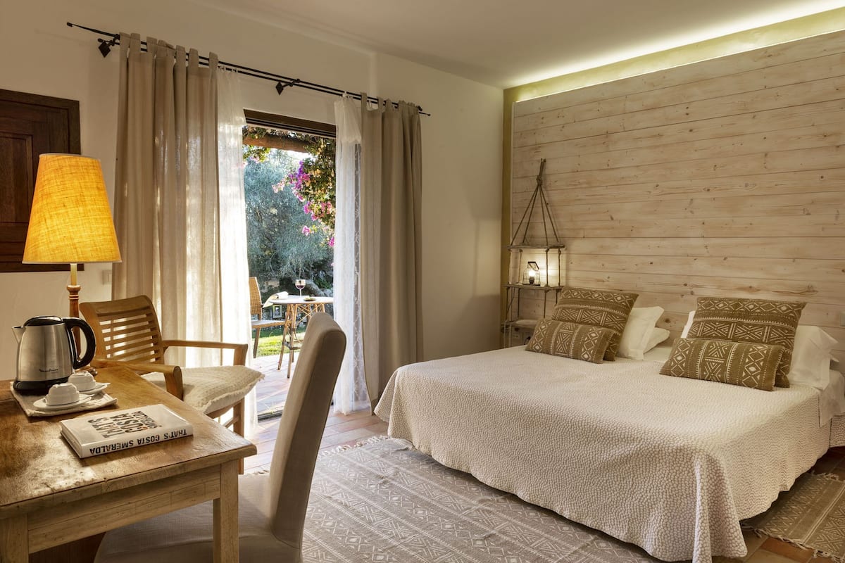 A bedroom with a wooden headboard, a bed with patterned pillows, a bedside table with a lamp, and a view of a garden through a window.