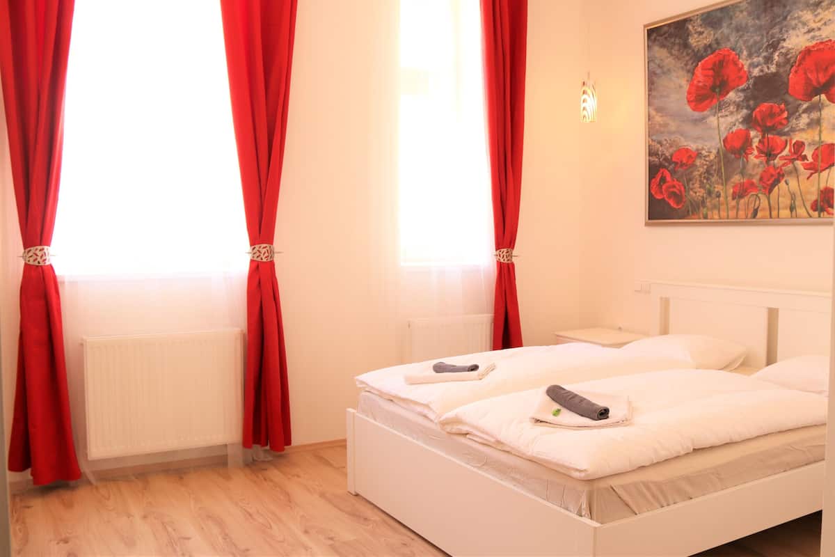 A bedroom with a white bed, red curtains, a radiator, and a painting of red flowers.
