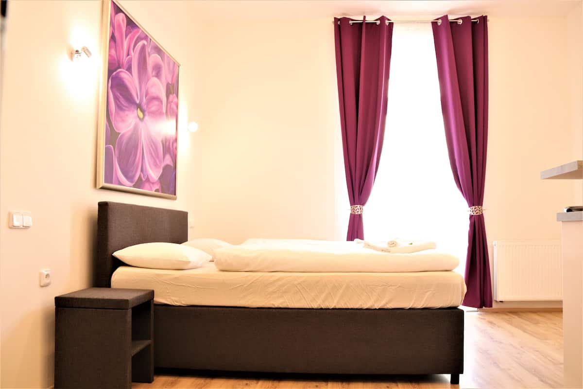 A bedroom with a bed, a nightstand, a wall-mounted light, and a window with purple curtains.