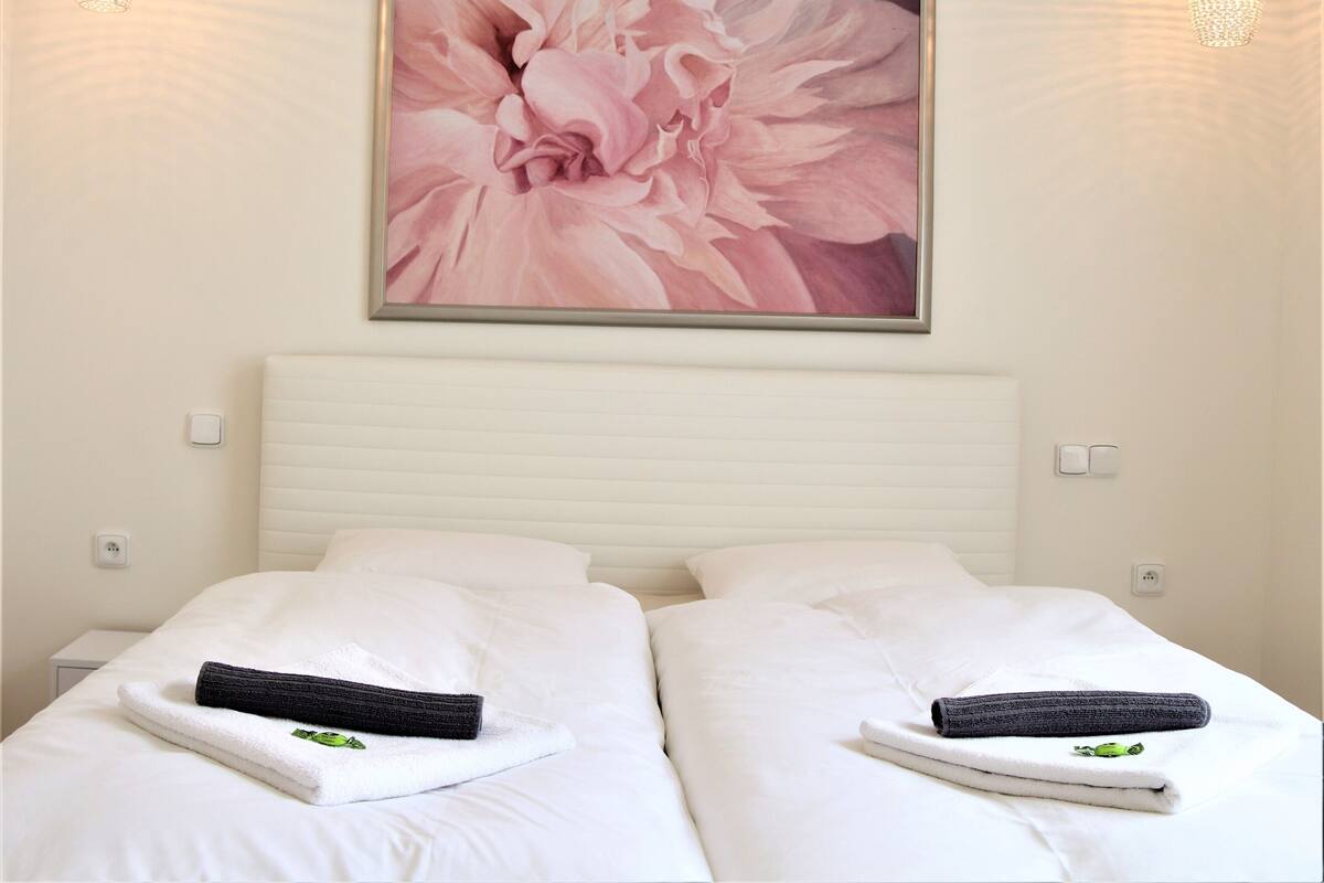 A hotel room with two beds, white bedding, a large floral artwork above the beds, and two pendant lights.