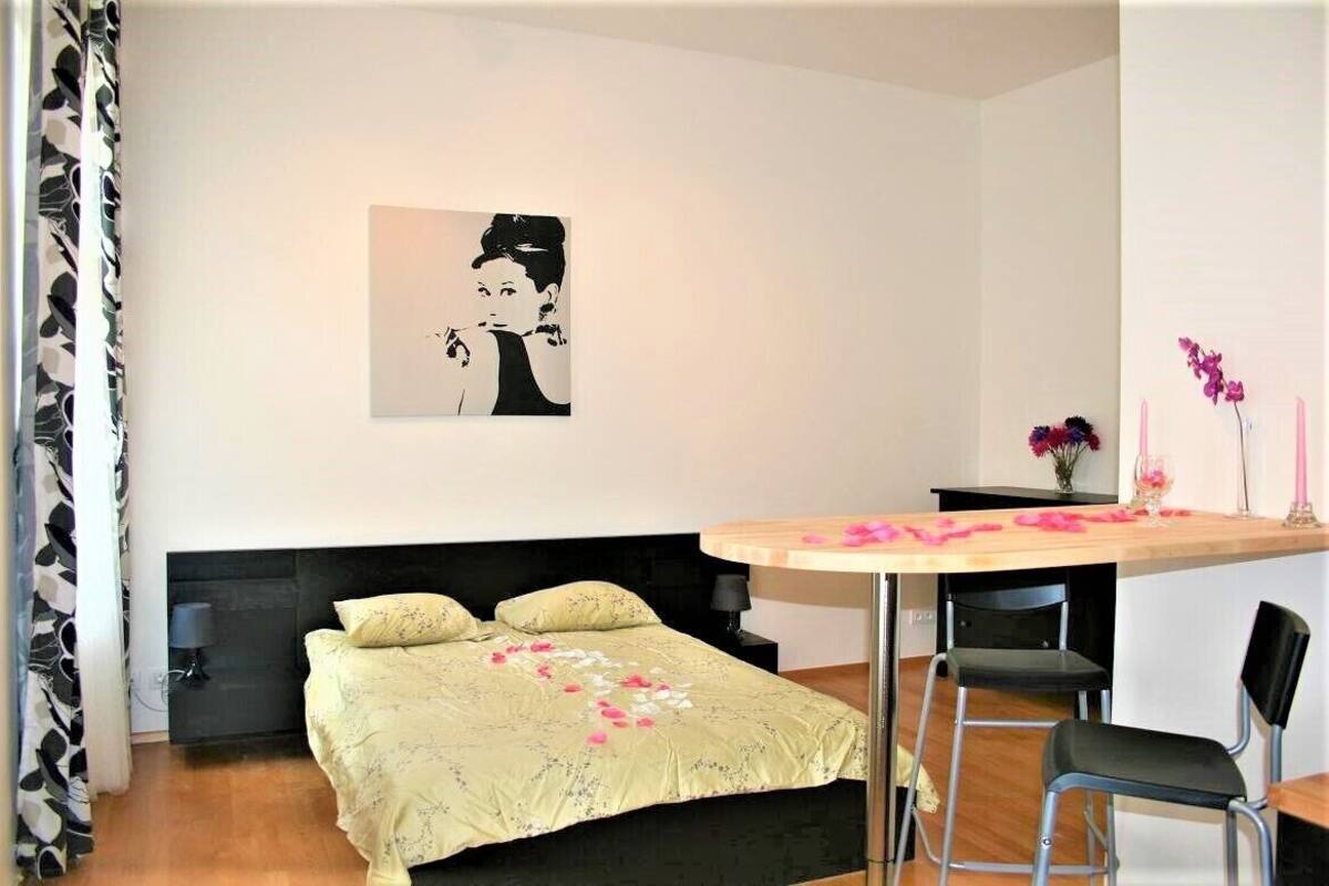 Une chambre à coucher comprenant un lit, une table de chevet, une chaise, une petite table avec un vase de fleurs et une œuvre d’art accrochée au mur.
