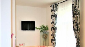 A small, well-lit room with a wooden desk, a chair, a bed with a floral bedspread, a television mounted on the wall, and a window with patterned curtains. - Reznicka Apartments (Prague)