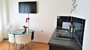 A modern kitchen with a black countertop, a built-in oven, and a sink. There is a dining area with a glass table and white chairs. A potted orchid is on the table, and a flat-screen TV is mounted on the wall. - Epstein Apartments (Prague)