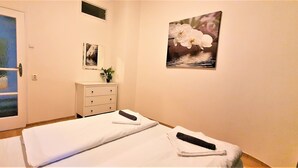 A hotel room with a bed, a bedside table, a small cabinet, a window with a view, and a wall-mounted picture. - Epstein Apartments (Prague)