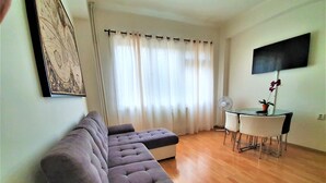 A living room with a grey sofa, a dining table with chairs, a flat-screen TV, and a potted plant. - Epstein Apartments (Prague)