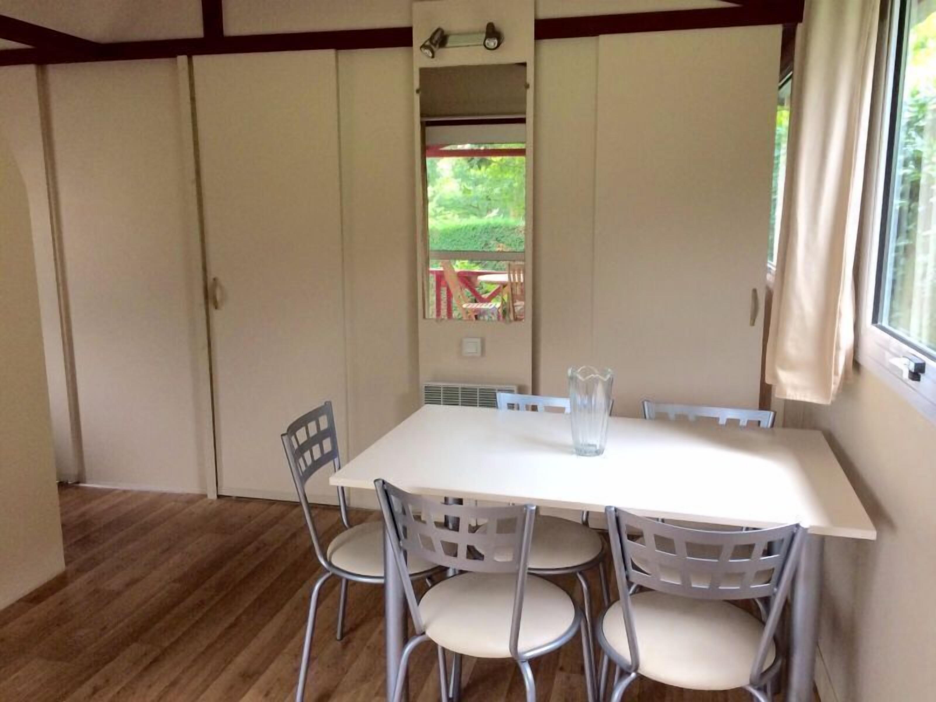 A dining area with a white table and four chairs, a vase on the table, and a window with a view of greenery outside.