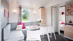 Una habitación moderna y bien iluminada con un sofá blanco, una mesa de comedor con sillas, un pequeño armario blanco y una ventana con vistas a zonas verdes.