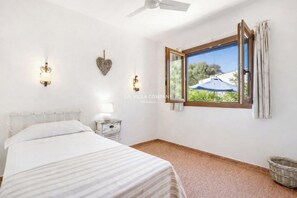 A bedroom with a bed, a nightstand, a wall-mounted light, a ceiling fan, and a window with a view of greenery.
