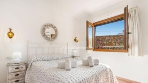 Quarto com cama, mesa de cabeceira, abajur, espelho e janela com vista para uma parede de pedra e flores.