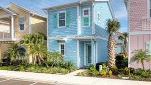 Une maison à deux étages, à l’extérieur bleu et aux volets verts, entourée de plantes tropicales et de palmiers.