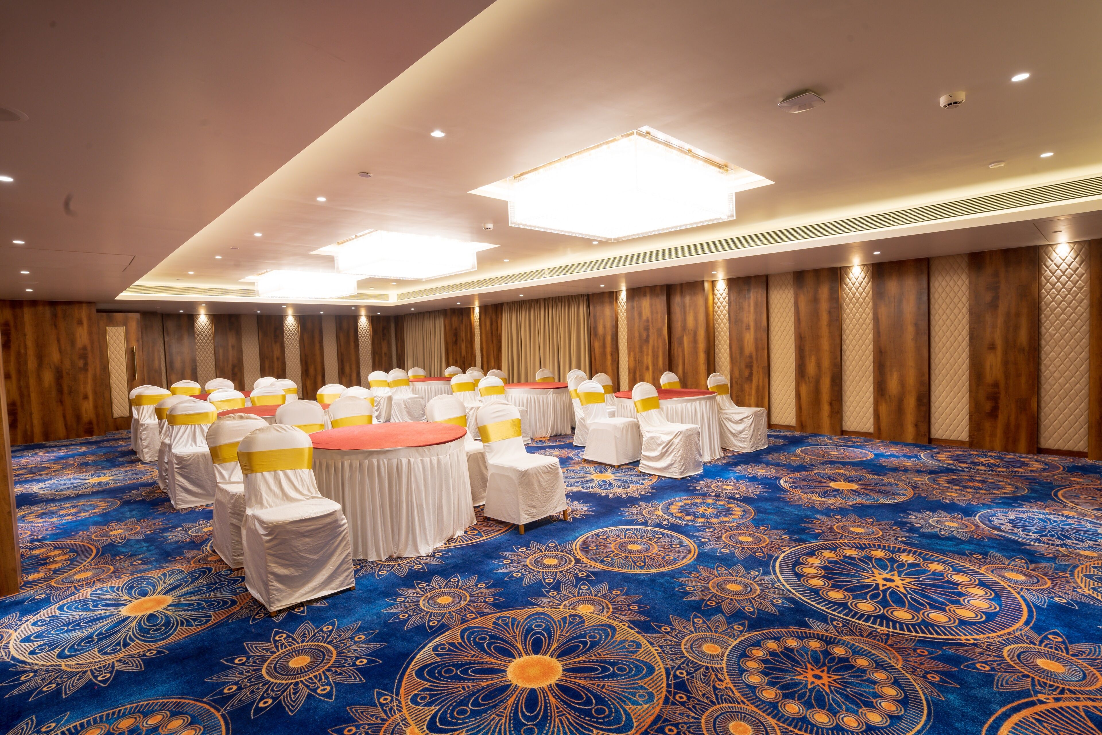 Une salle de conférence avec des tables rondes, des chaises blanches ornées de rubans jaunes et un tapis bleu à motifs.
