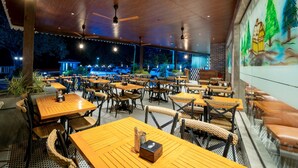 Un área de comedor al aire libre con mesas y sillas de madera, ventiladores de techo y un mural en la pared.