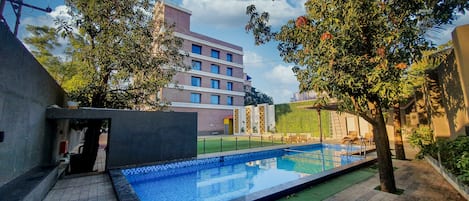 Una piscina con borde de baldosas, rodeada de árboles y un edificio al fondo.