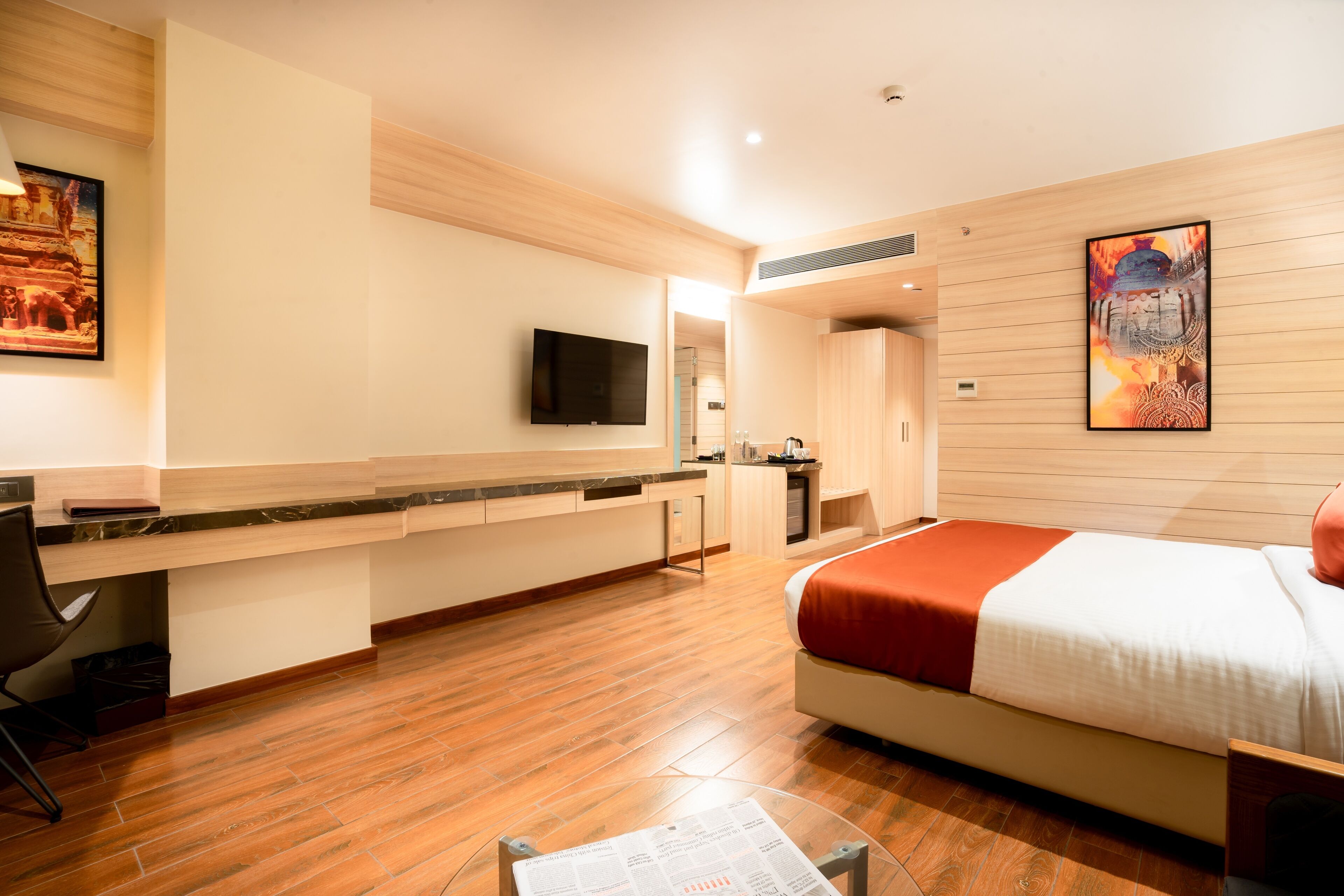 Een moderne hotelkamer met een houten vloer, een bed met een rode deken, een bureau met een stoel, een flatscreen televisie en kunst aan de muren.