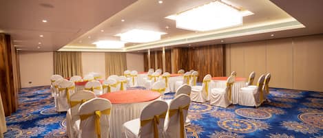 Un salón de conferencias con mesas redondas y sillas blancas, suelo alfombrado e iluminación en el techo.