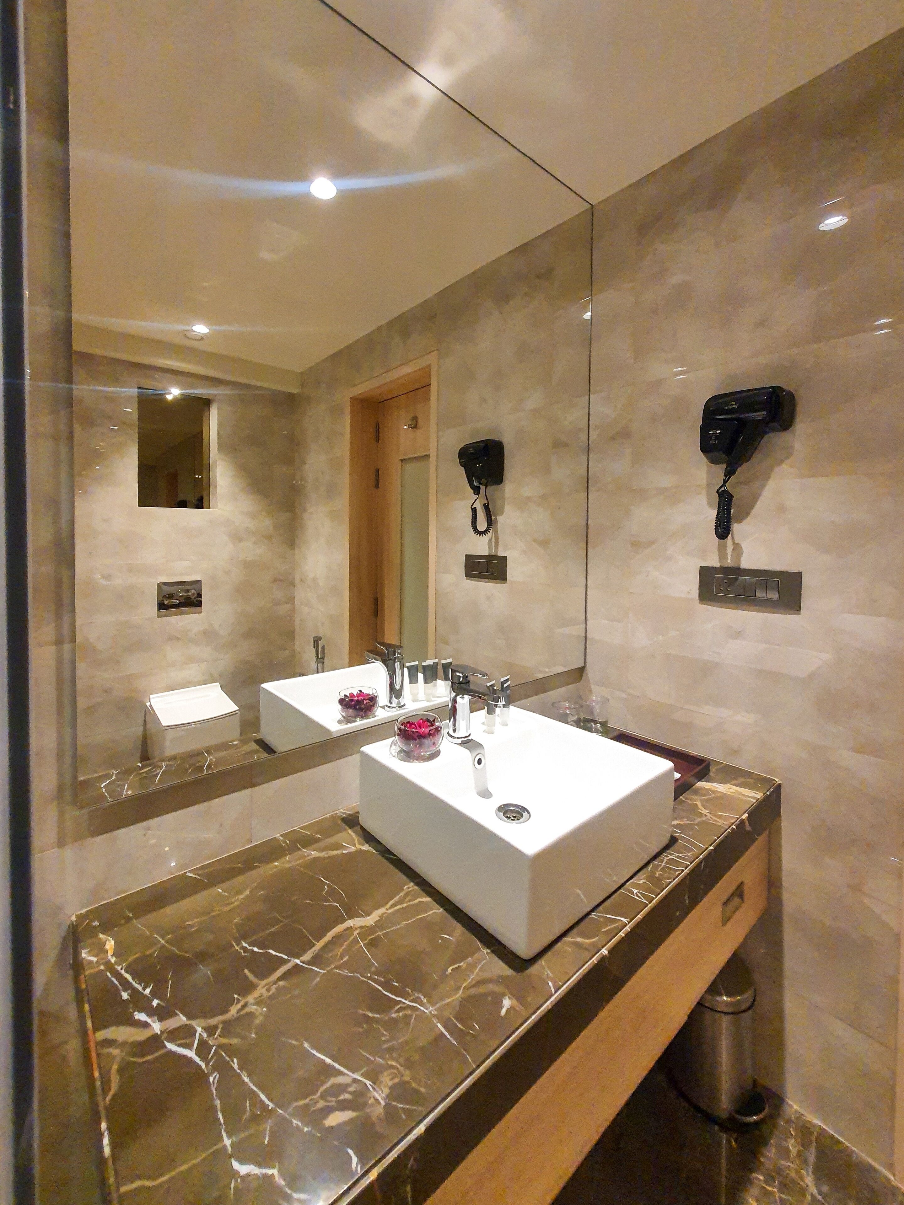 Un baño moderno con un espejo grande, un lavabo blanco, encimera de mármol, secador de pelo montado en la pared y un cubo de basura.