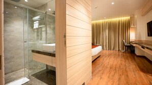 Una habitación de hotel moderna con cabina de ducha de vidrio, tocador de madera, cama con cabecero y una ventana grande con cortinas.