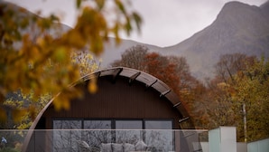 A modern building with a curved roof and large windows, surrounded by autumn foliage and a mountain in the background. - SeaBeds Luxury Lookouts with Hot Tubs (Ballachulish)
