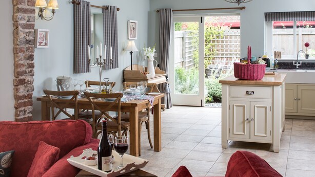 A cozy living room with a red sofa, a dining area with a wooden table and chairs, and a kitchen with a white cabinet and a red basket. - The Dovecote Barns (York)
