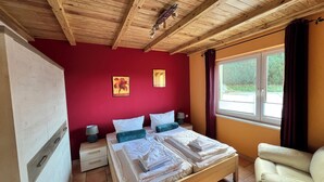 Un dormitorio con techo de madera, paredes rojas, cama con ropa de cama blanca, dos almohadas, una mesita de noche, una lámpara y una ventana con cortinas.