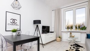 Ein modernes Wohnzimmer mit einem schwarzen Esstisch, klaren Stühlen, einem weißen Sofa, einem Fernseher, einer Lampe und einem großen Fenster mit Pflanzen.