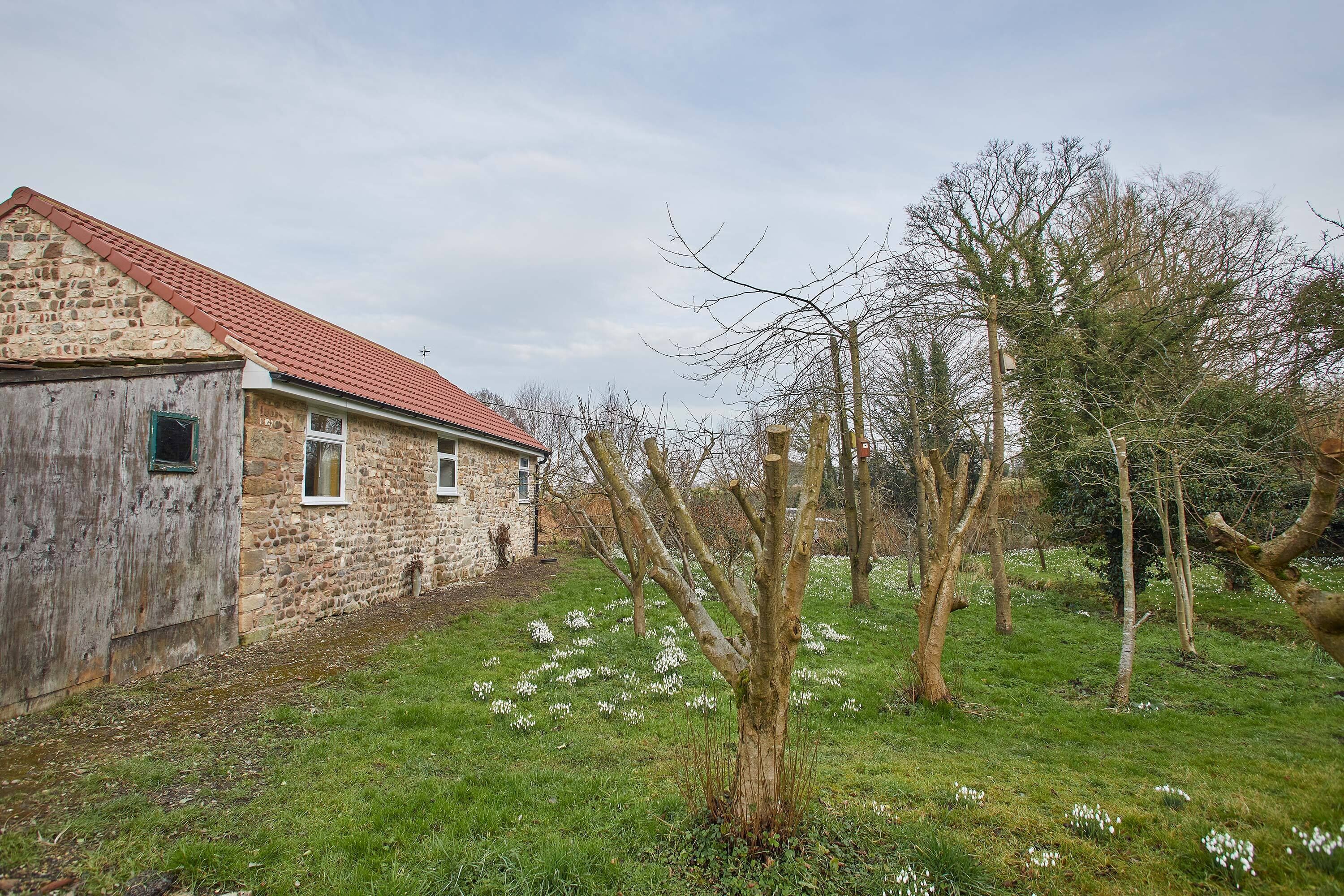 Un bâtiment en pierre avec un toit rouge, entouré d’une zone verdoyante parsemée d’arbres et de fleurs blanches.