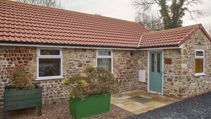 A stone building with a red-tiled roof, a green door, and a small garden area with a bench and potted plants. - Snowdrop Cottage (Knaresborough)