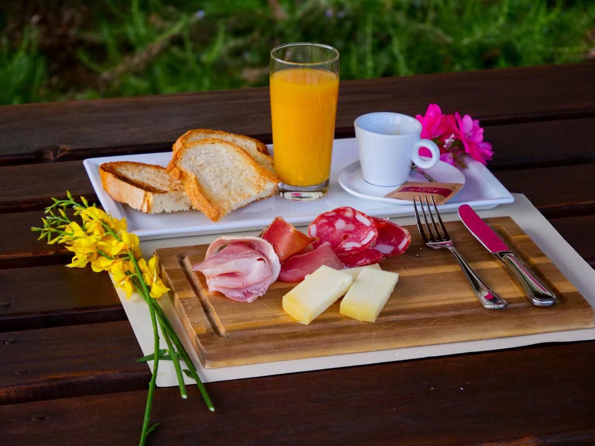A picnic setup with bread, cheese, ham, and salami on a wooden board, a glass of orange juice, a cup, and cutlery on a white tray.